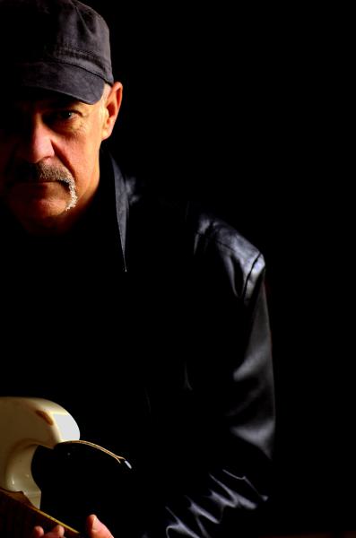 Bandfoto Wolfgang Scheele Band: Porträt eines Gitarristen  Ein Mann steht halb im Schatten vor schwarzem Hintergrund. Er trägt eine dunkle Schirmmütze und eine schwarze Lederjacke. Sein Gesicht ist nur von der linken Seite beleuchtet, wodurch ein starker 